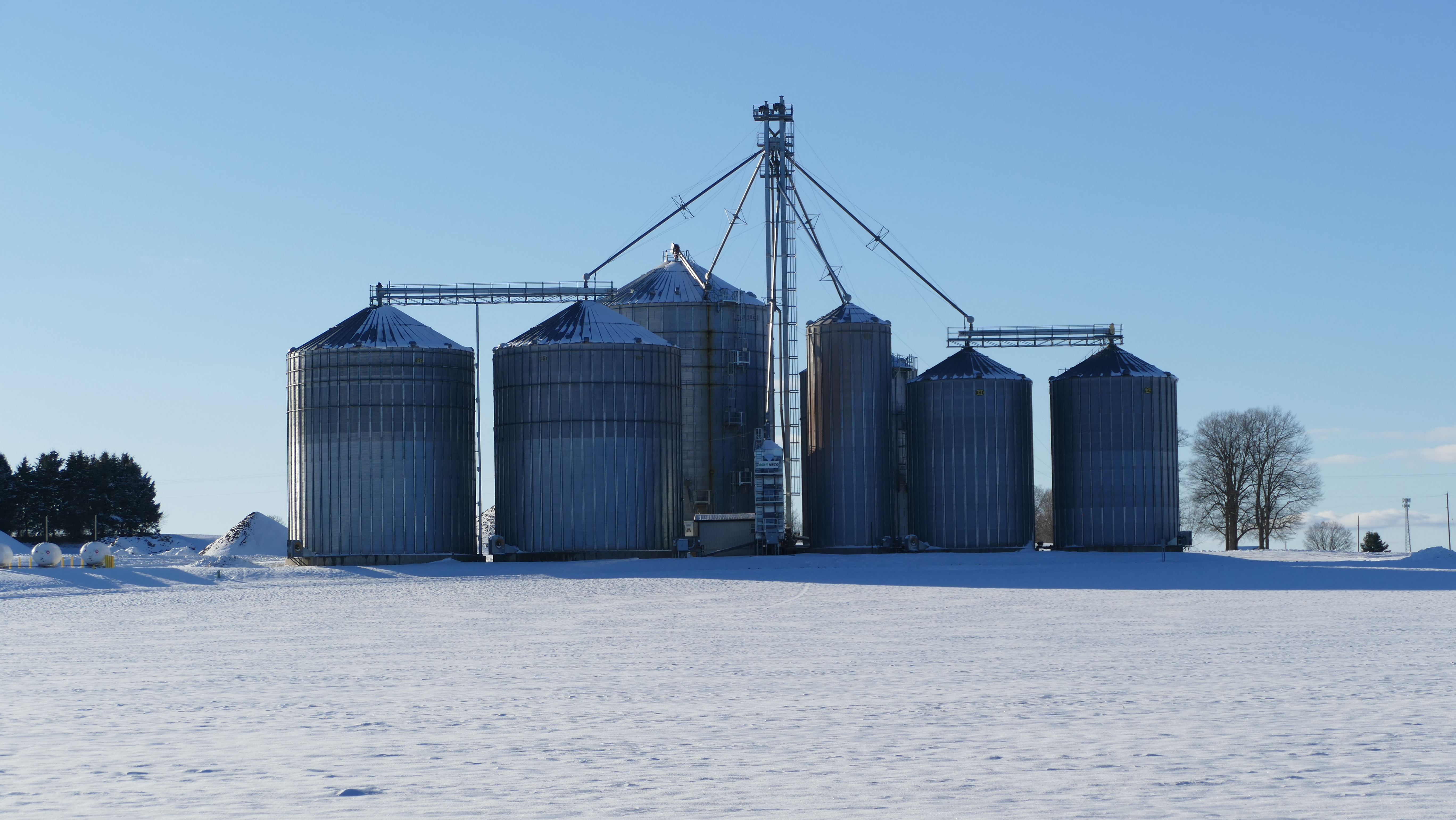 Several large metal grain bins and silos stand in a row on a farm, surrounded by snow-covered ground.
